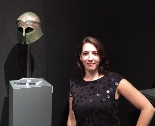 Picture of a woman in black dress in front of a bronze ancient Greece helmet.
