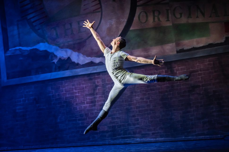 An image of a male ballet dancer in a white costume executing a grand jeté with arms outstretched, leaping across a stage with a brick wall and faded signage in the background.