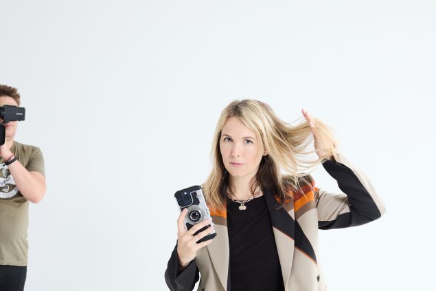 A blonde woman in a geometric-patterned jacket holds a smartphone with a vintage camera case in one hand while lifting her hair with the other, looking directly at the camera, as a man in the background wearing a t-shirt and bracelets holds a Sony video camera up to his face.