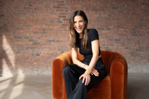 A smiling woman in a black top and pants sits on an orange velvet chair in front of a red brick wall, basking in natural light.