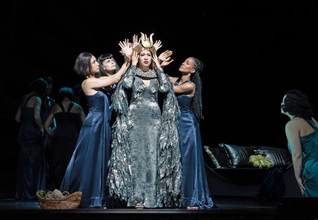 A group of women in formal blue gowns surrounds a central figure in a silver costume and feathered cape, placing a gold headdress on her during a stage performance.