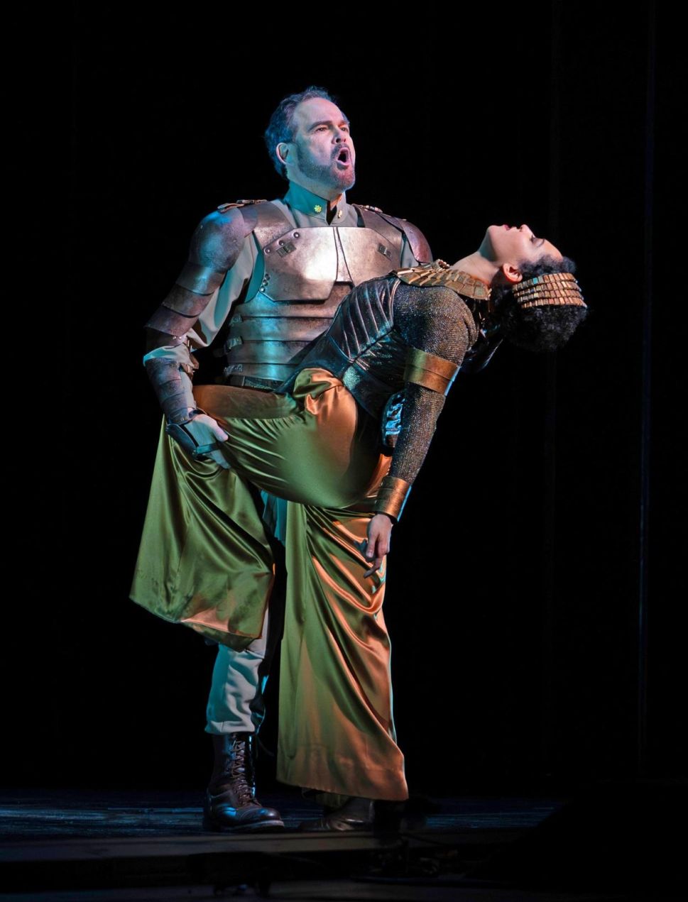 A man in armor holds a woman in gold and black costume, who is leaning back dramatically with her eyes closed, during an intense stage moment.