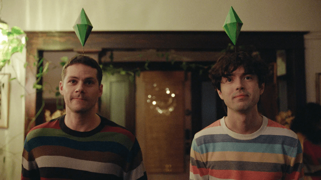 A man in a striped sweater stands beside another man in a differently striped sweater, both wearing green diamond-shaped headpieces, during a scene from Twinless.